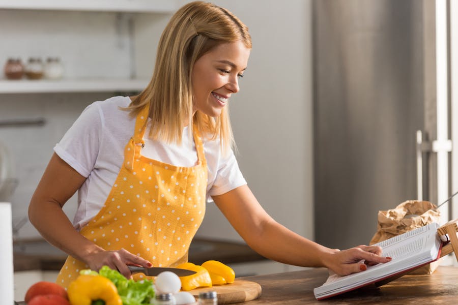 Smiling young woman in apron reading cookbook while cooking in kitchen